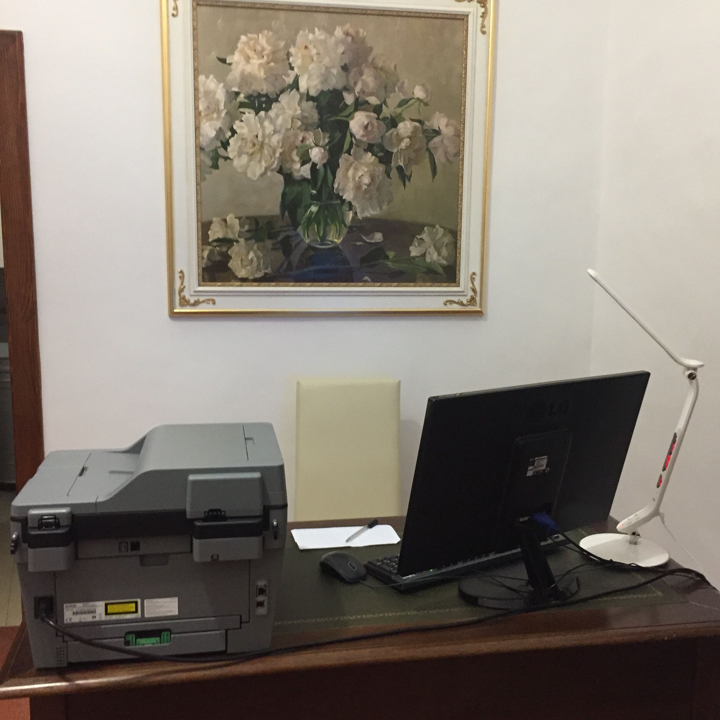 A desk with a printer, a laptop, and a computer monitor. There is a framed painting of flowers on the wall above the desk.