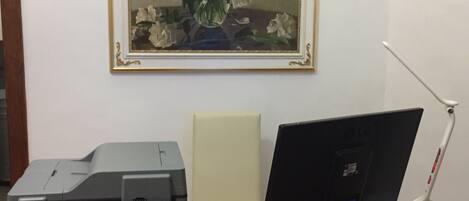 A desk with a printer, a laptop, and a computer monitor. There is a framed painting of flowers on the wall above the desk.