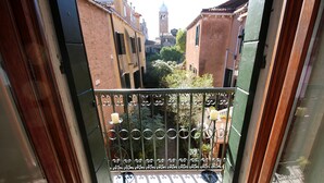 A balcony with a wrought iron railing, a view of a narrow street with buildings, and a tower in the distance. - SUNNY Ghetto canal (Venezia)