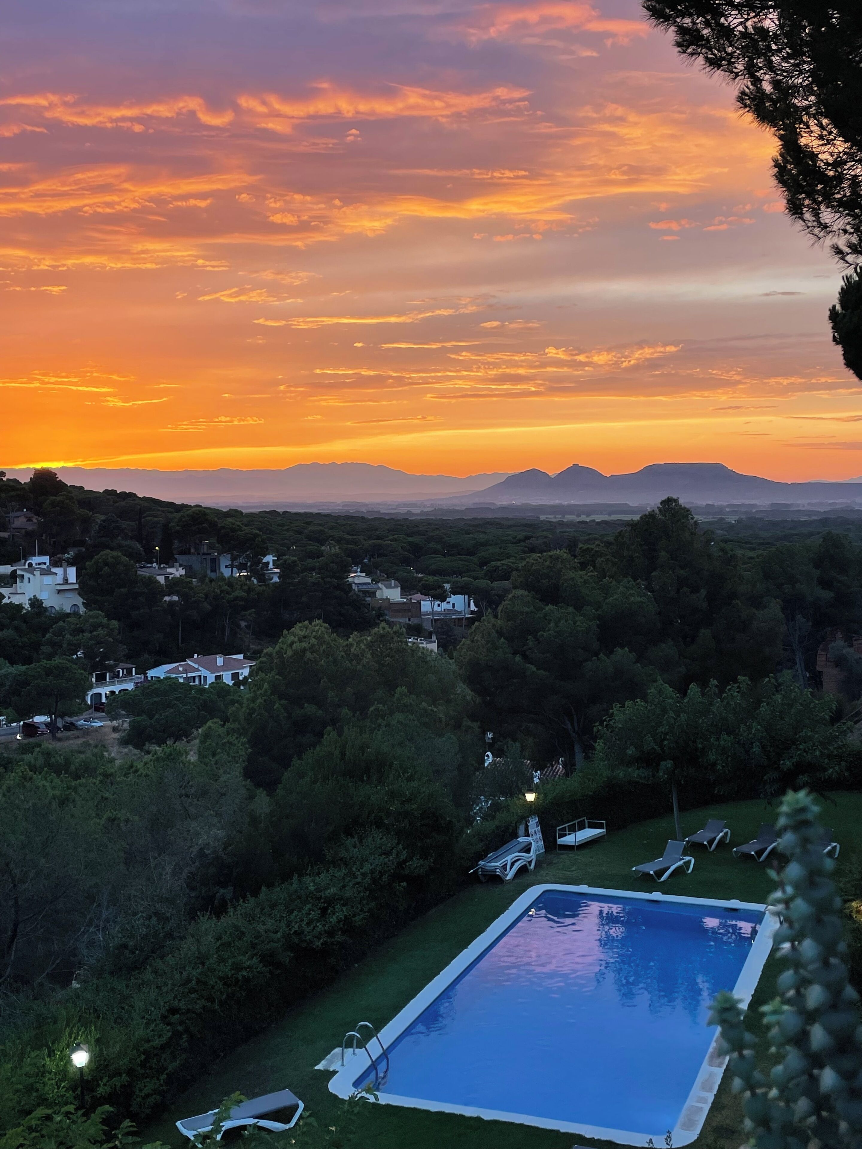 A swimming pool surrounded by lush greenery, with a view of distant mountains and a sunset sky.