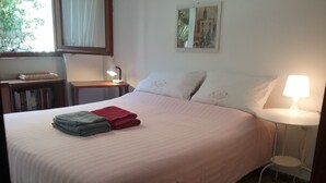 A neatly made bed with folded towels, a bedside table with a lamp, a small chair, and a bookshelf. - Apartment with sea view, 5 minutes from the beach (Finale Ligure)