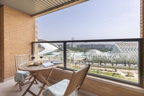 Een balkon met een houten tafel en stoelen, uitzicht op een modern gebouw en een heldere lucht.