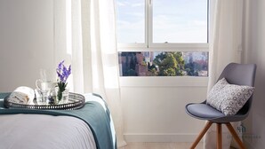 A neatly made bed with a teal bedspread, a tray with a towel, glasses, and a vase with flowers, and a chair with a patterned pillow by a window with a city view. - Alcazaba Exclusive View Living4malaga (Málaga)