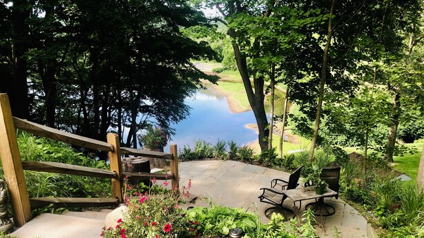 A wooden deck with a stone pathway leading to a seating area with a table and chairs, surrounded by lush greenery and a view of a lake. - Moonriver Inn (Narrowsburg)