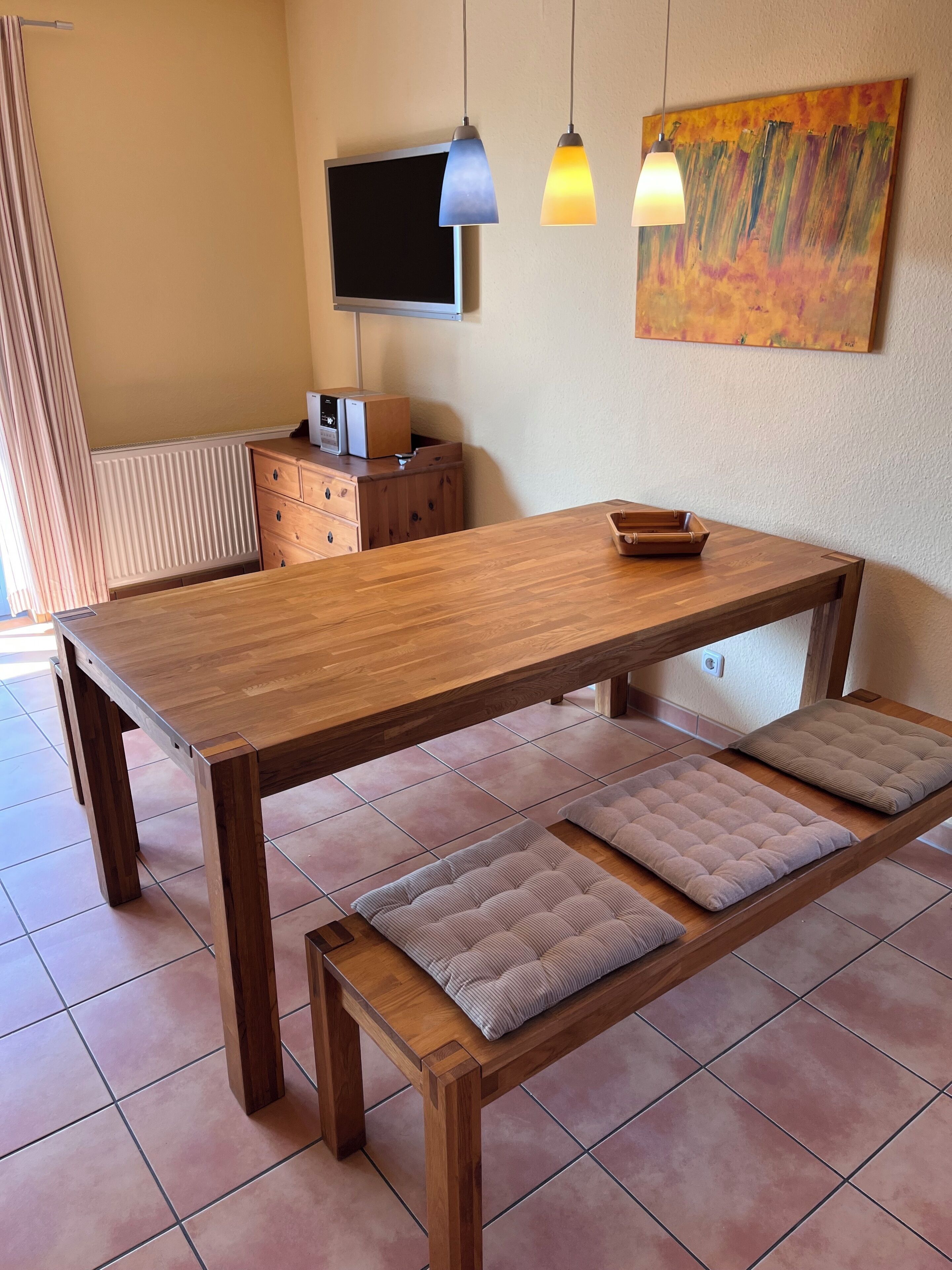 Ein Esszimmertisch aus Holz mit Bänken, einem Fernseher und Pendelleuchten in einem Raum mit gefliestem Boden.