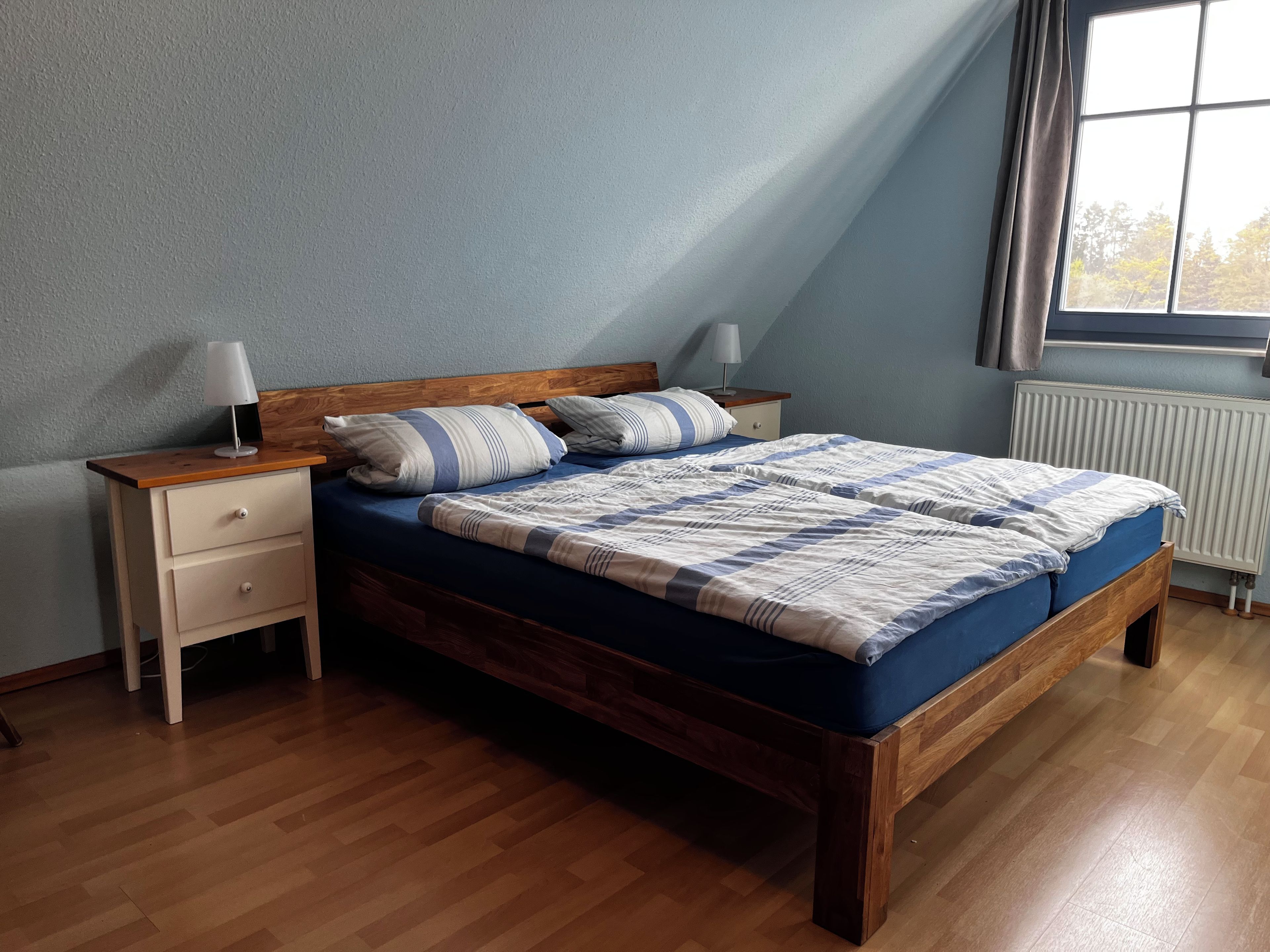 Een slaapkamer met een houten bed, een nachtkastje, een lade, een lamp en een raam met gordijnen.