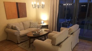 Un salon comprenant un canapé, un fauteuil et une table basse. On y trouve une lampe sur une table d’appoint et un lustre au plafond.