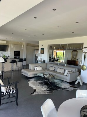 A modern living room with a sectional sofa, a glass coffee table, and a unique rug. There is a dining area with a bar and high chairs. The kitchen is visible in the background.