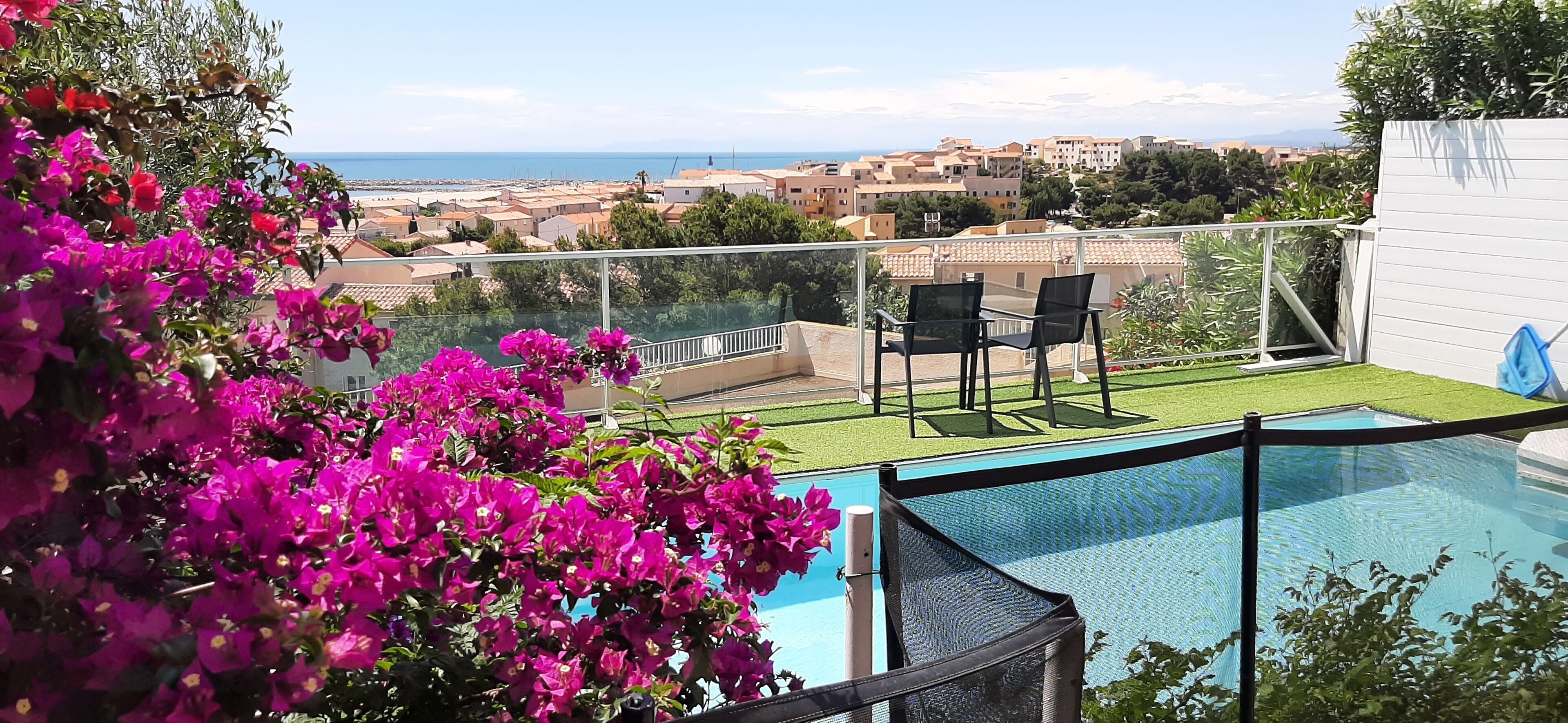 A poolside area with a view of the sea, a grassy lawn, and a seating area with two chairs.