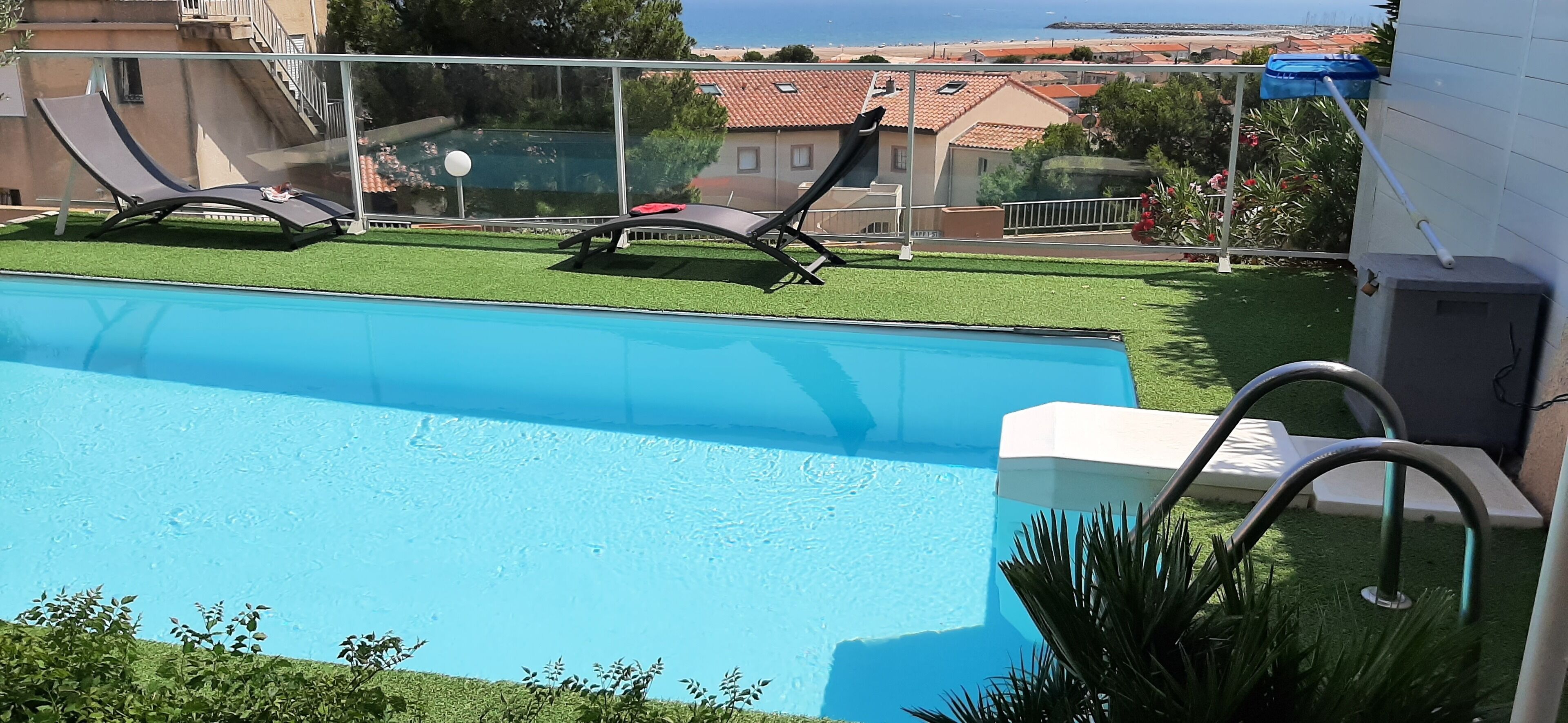 Una piscina con dos sillas reclinables, un pequeño jardín con césped artificial y vistas a una zona residencial con casas y un cuerpo de agua al fondo.
