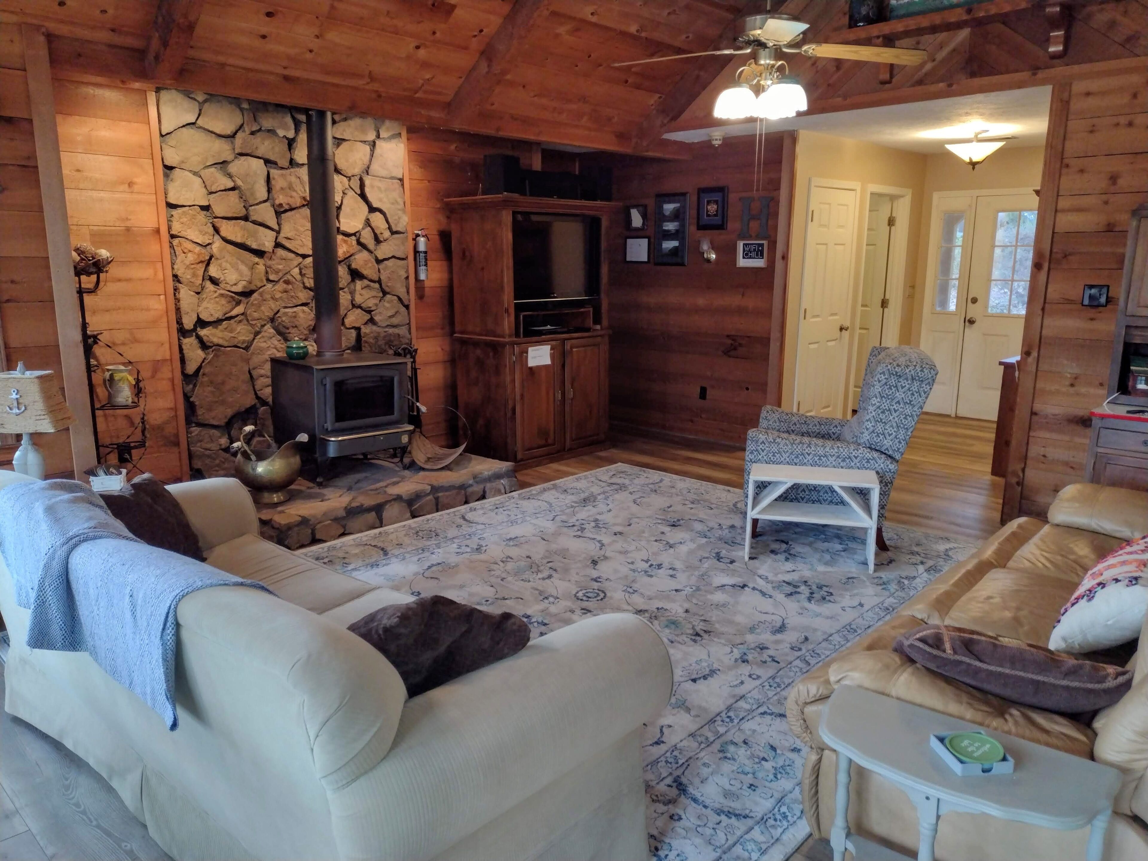 Interior acogedor de una cabaña con pared de piedra, muebles de madera, chimenea y un ventilador de techo.