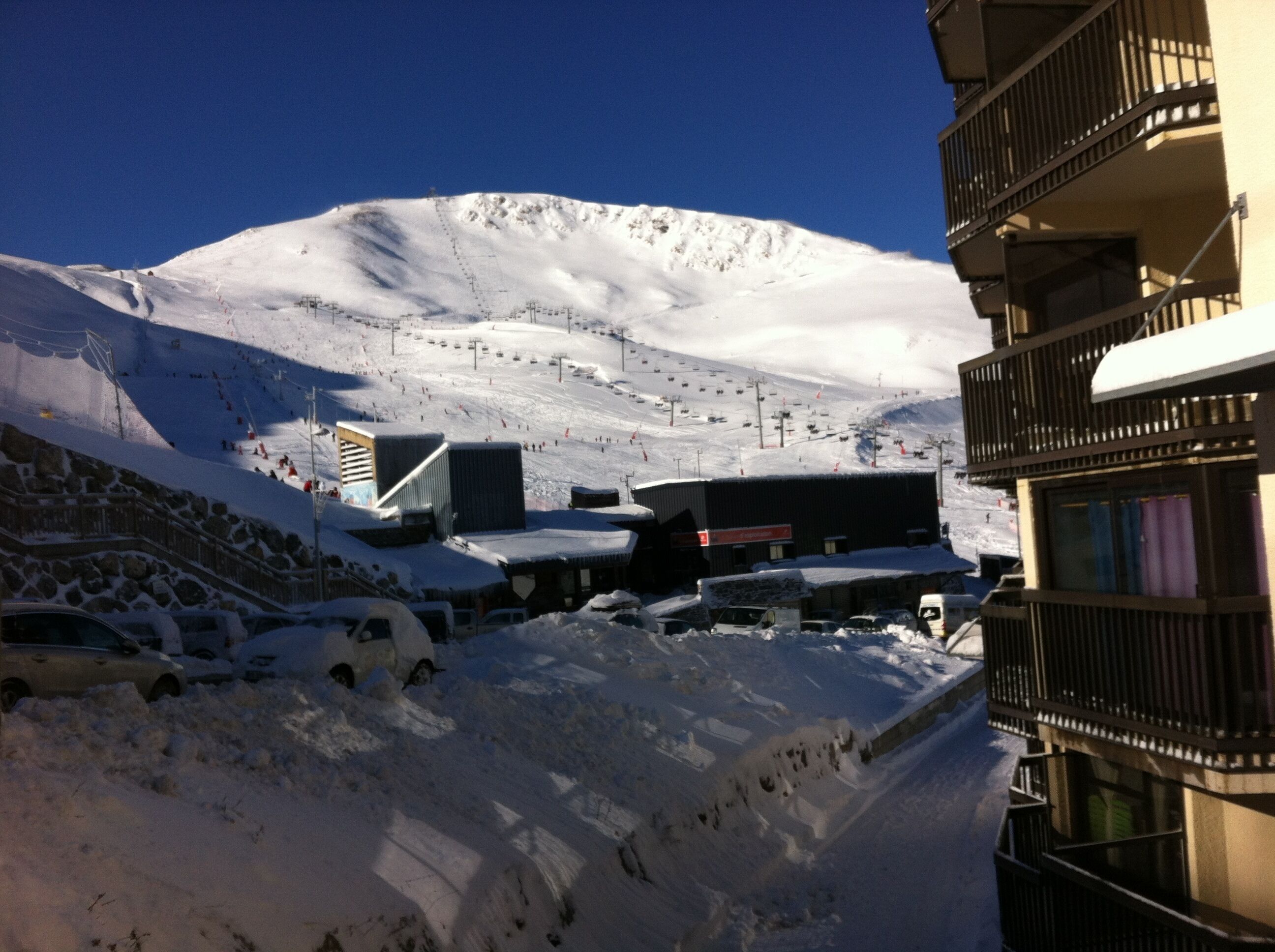 Une montagne enneigée avec un télésiège, un bâtiment avec une enseigne et un parking avec des voitures.