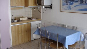 A compact kitchen with wooden cabinets, a microwave, and a sink. There is a dining table with chairs and a painting of a snowy landscape on the wall.
