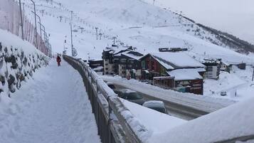 Une route de montagne enneigée, bordée de bâtiments, dont un bar et un restaurant, avec un télésiège visible au loin.