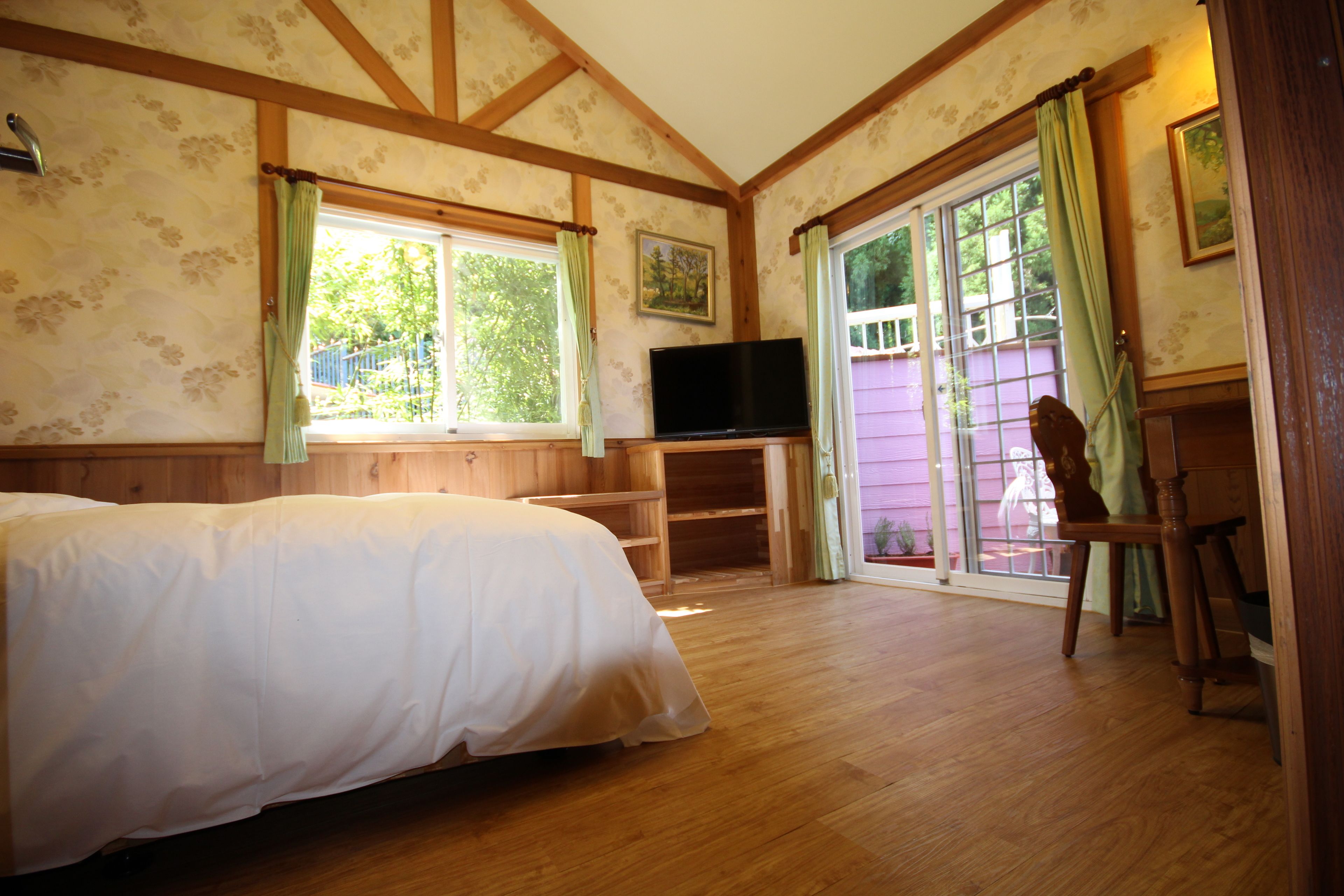 Un dormitorio con cama, televisor, una silla de madera y un ventanal corredero que da a un espacio exterior.