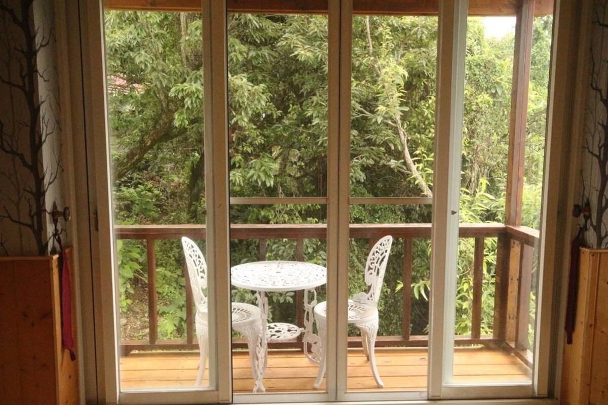 Una terraza de madera con una mesa y sillas blancas, con vista a un bosque frondoso.