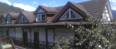 A row of chalet-style buildings with pitched roofs and dormer windows, a wooden balcony, and a view of a mountainous landscape.