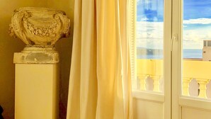 A room with a large window, a white curtain, and a stone urn on a pedestal. - Flat in Beausoleil, Monaco: double glazing, terrace, sea view & paying parking (Beausoleil)