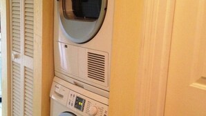 A front-loading washing machine and dryer stacked in a laundry room with a door and a window with blinds. - Park Shore Resort #355 Lake view, Tennis, Bike/Walk, Gym, Beach, Pickleball (Naples)