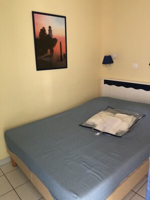 Una cama individual con colchón gris y almohada, una lámpara de pared y un cuadro con una imagen de un atardecer.