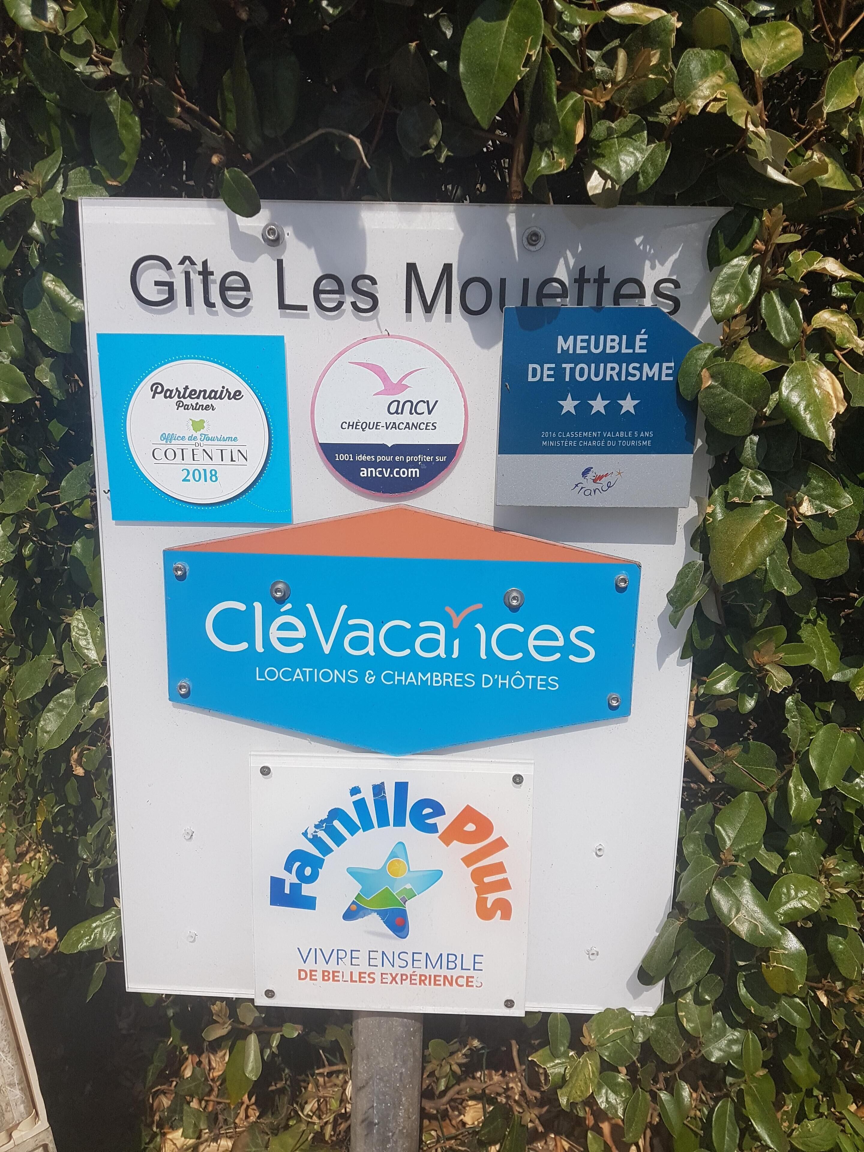 A sign with multiple logos and text, including 'Gîte Les Mouettes', 'Partenaire Office de Tourisme Cotentin 2018', 'ancv', 'CleVacances', and 'FamillePlus'.