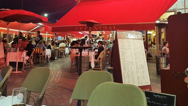 Ein Essbereich im Freien mit Tischen und Stühlen unter roten Sonnenschirmen, ein Schild mit der Aufschrift 'Brasserie Le Central' und eine Speisekarte.