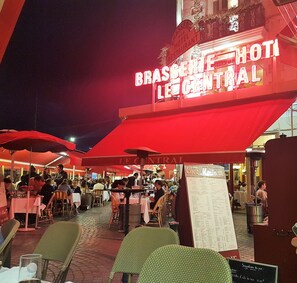 Un área de comedor al aire libre con mesas y sillas bajo sombrillas rojas, un letrero de 'Brasserie Le Central' y un cartel de menú.
