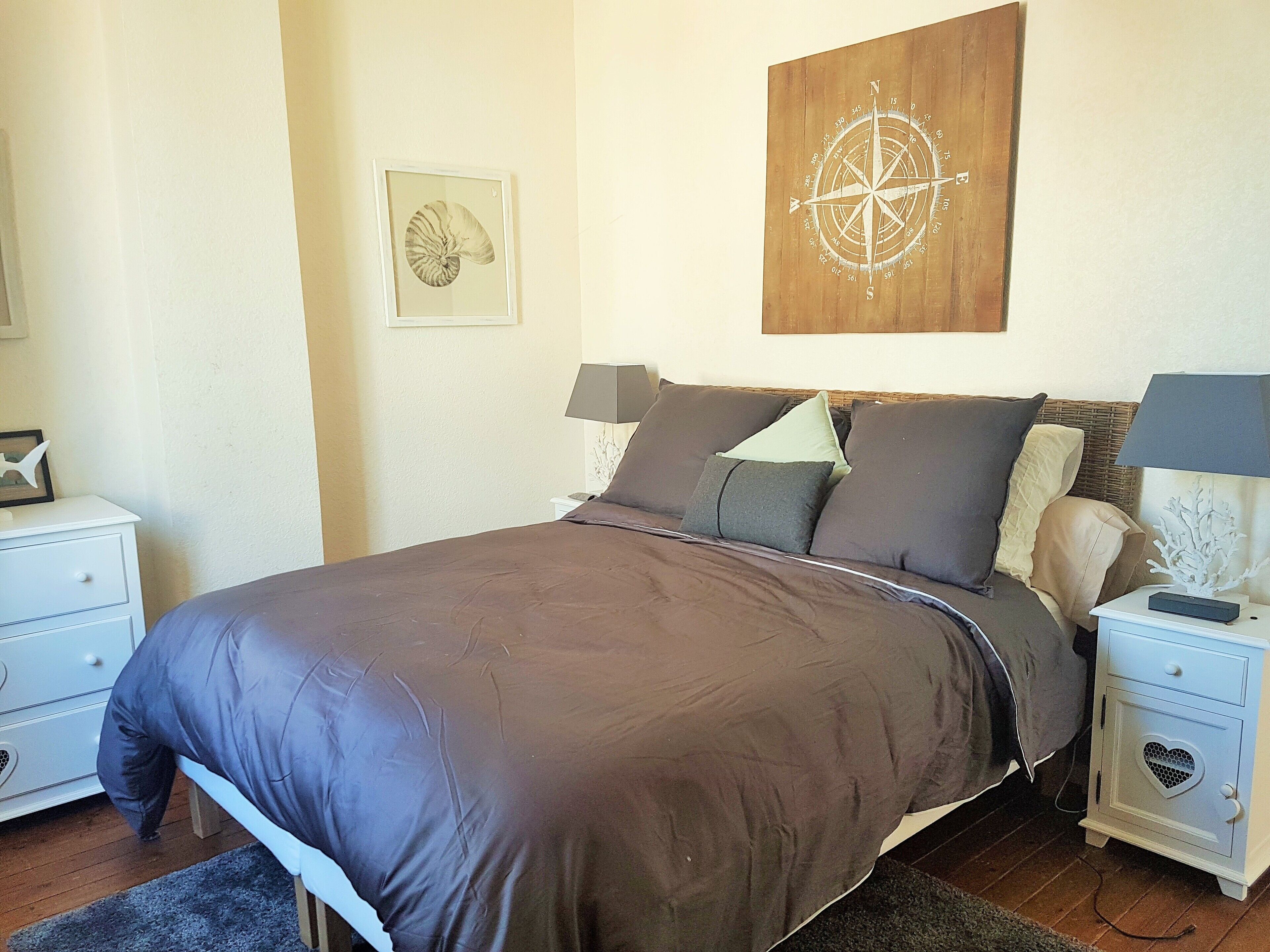 A neatly made bed with a brown bedspread, two bedside tables with lamps, a wall clock, and a framed compass artwork.