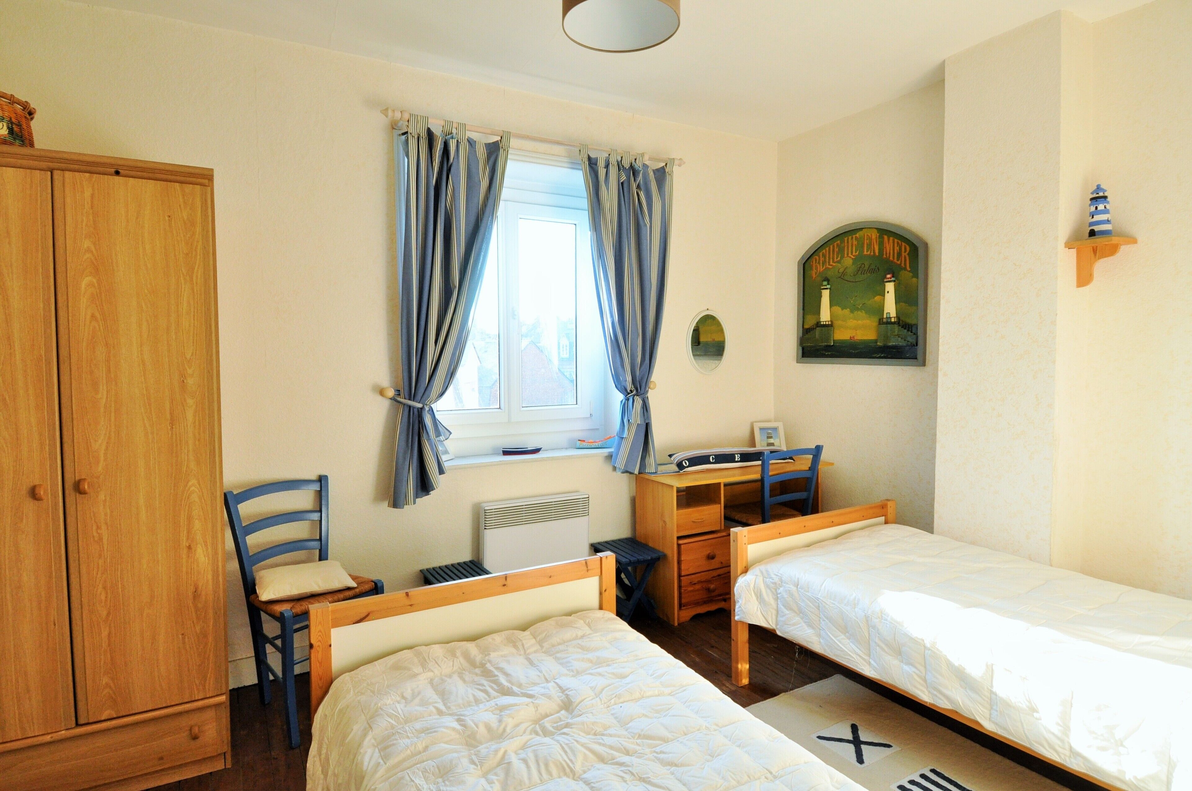 A bedroom with two single beds, a wooden wardrobe, a desk with a chair, a radiator, and a window with curtains.