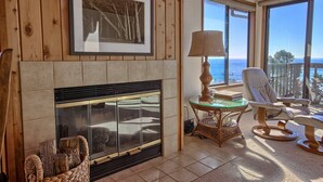 A cozy room with a fireplace, a large window with a view of the ocean, a comfortable chair, and a table with a lamp. - Panoramic Views From This Monthly Rental in Cambria! Stunning! (Cambria)
