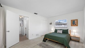 A bedroom with a bed, bedside lamps, a window with blinds, a door, and a hallway leading to another room. - Spacious 3 Bedroom 2 Bath Walk to Eagle Lake Park Fenced Yard (Clearwater)