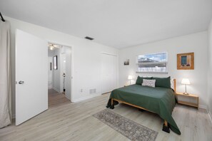 Un dormitorio con cama, lámparas de noche, ventana con persianas, una puerta y un pasillo que lleva a otra habitación.