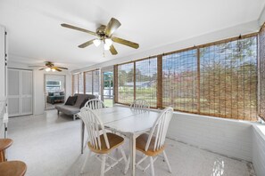 Un comedor con mesa y sillas, ventilador de techo y vistas a través de una ventana con celosía de bambú.