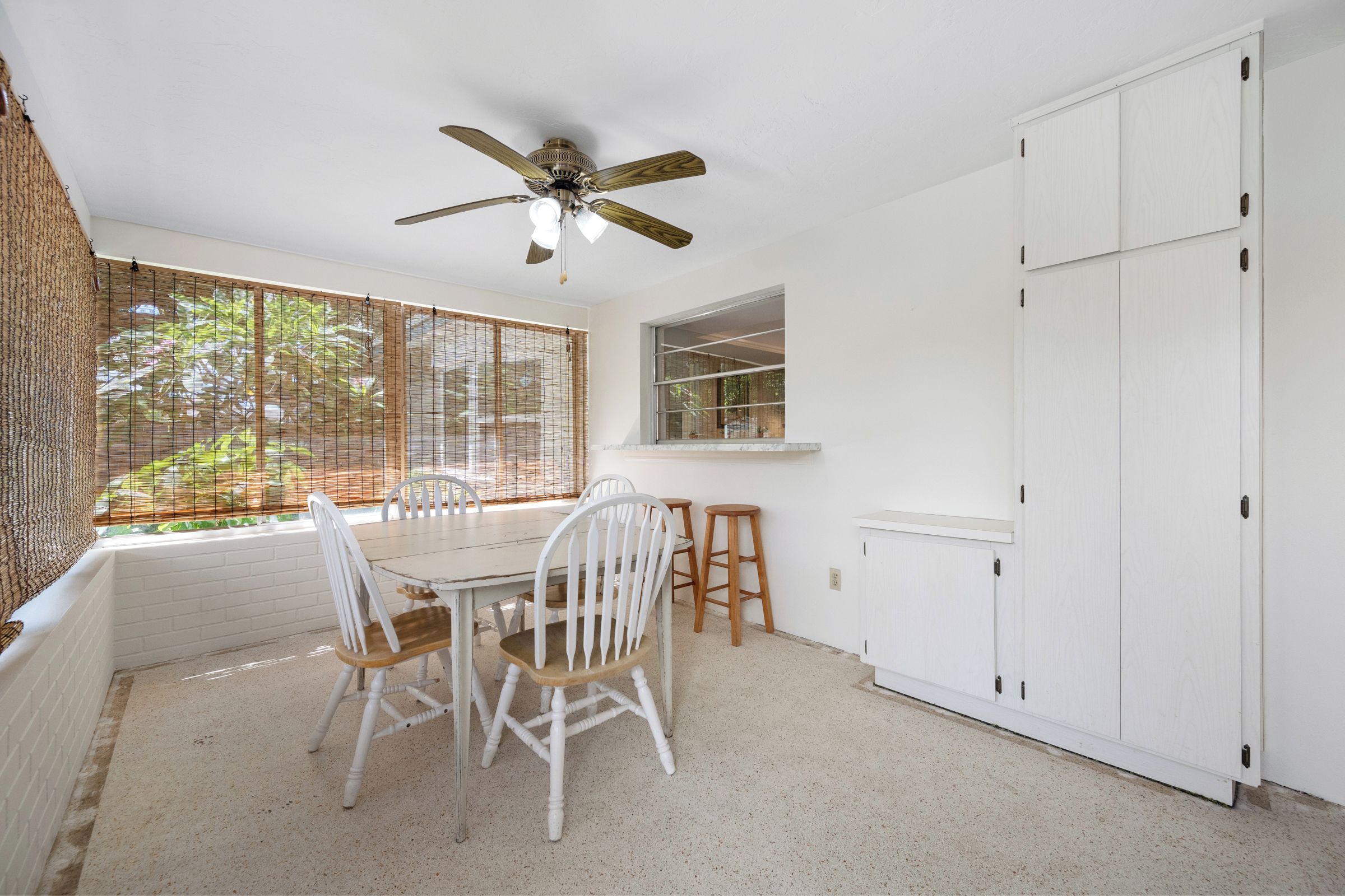 Quarto com mesa de jantar, cadeiras, ventoinha no teto, janela com persianas e guarda-roupa branco.