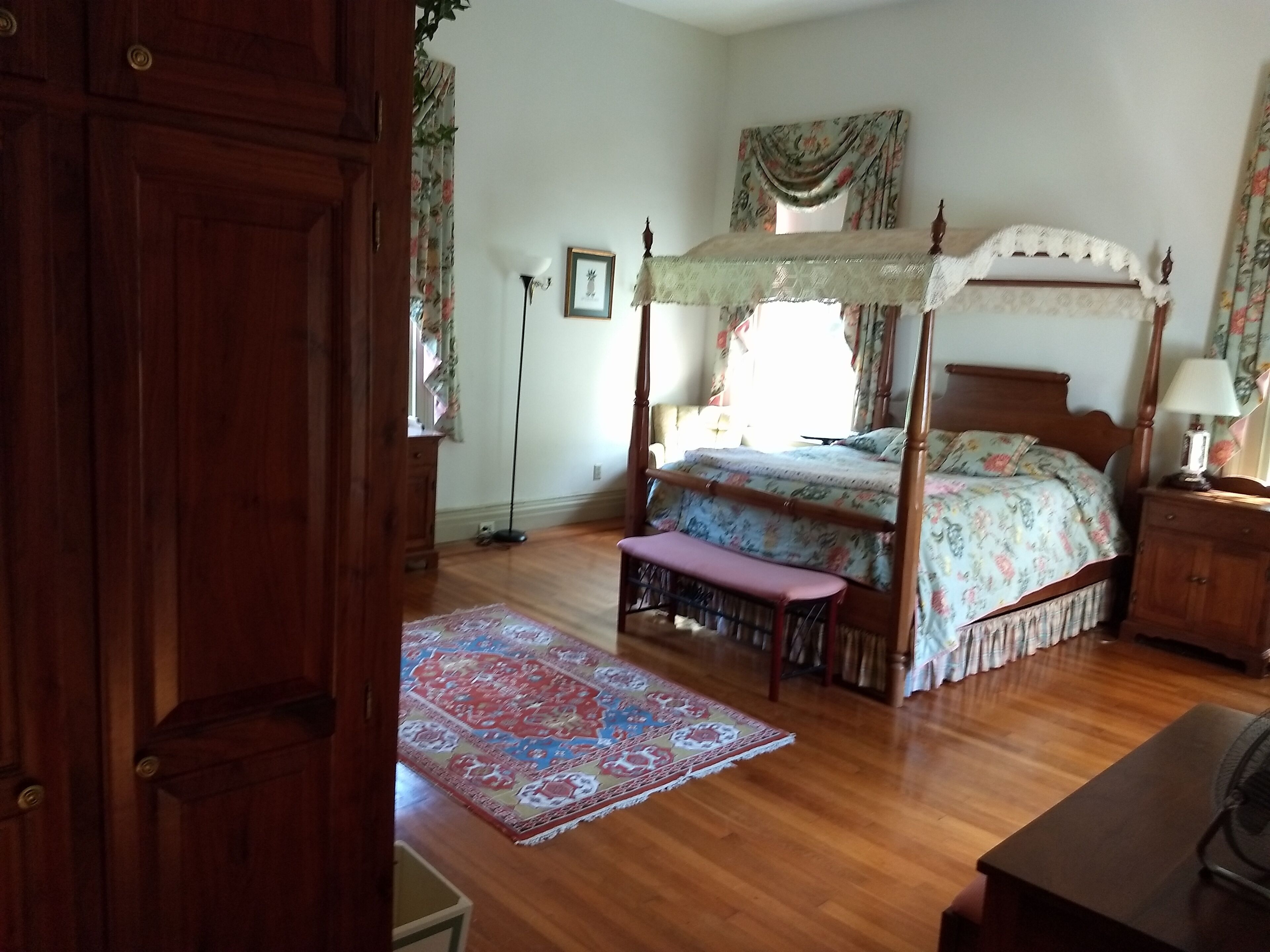 A traditional four-poster bed with floral bedding, a wooden bench, and a red patterned rug in a well-lit room with wooden flooring.