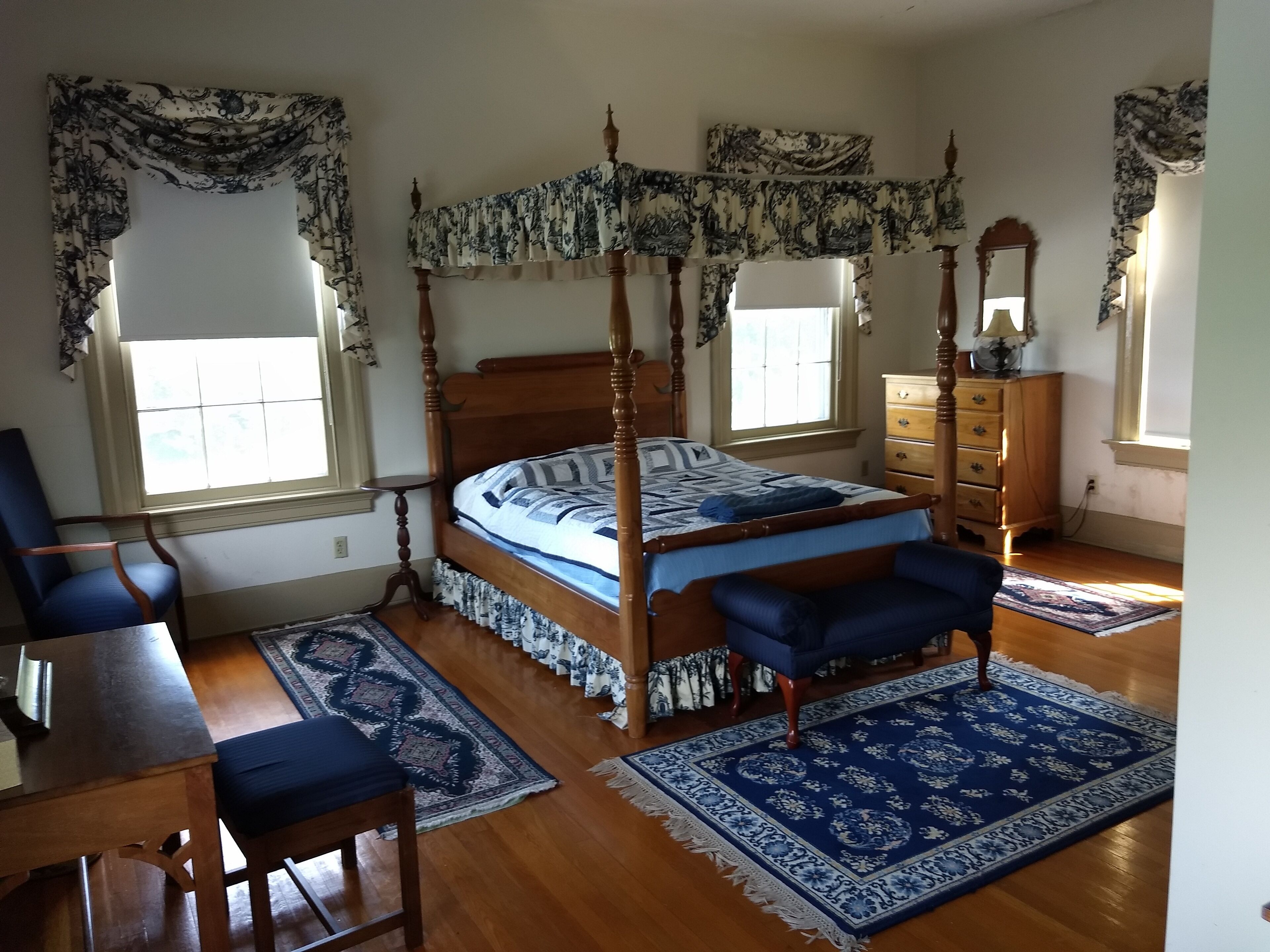 A four-poster bed with a floral bedspread, a wooden dresser, a blue chair, and a blue rug in a bedroom with two windows and a mirror.