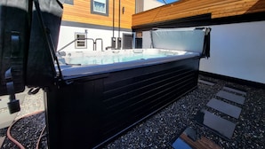 Een bubbelbad in een achtertuin met een houten terras, een zwembad en een huis op de achtergrond.