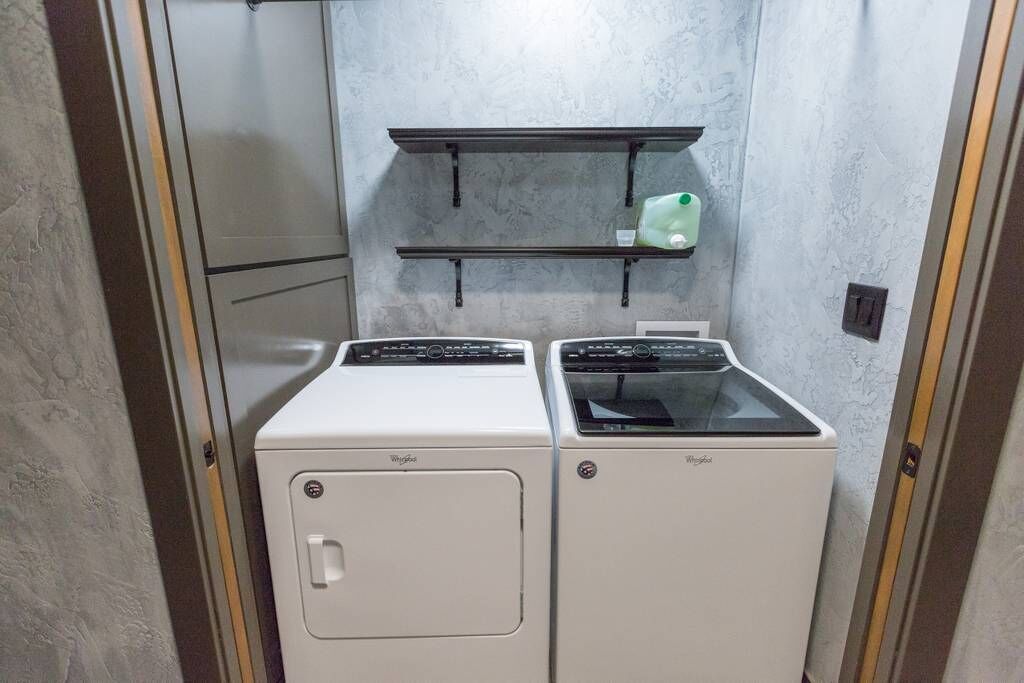 Lavandería con dos lavadoras y secadoras Whirlpool, un estante con una botella verde y una pared gris con textura.