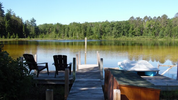 Un muelle de madera que lleva a un lago tranquilo, con dos sillas Adirondack y un bote cubierto al final.