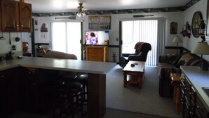 Cocina amplia con una isla central, taburetes de bar y un área de comedor con mesa y sillas. Se observa un televisor al fondo, y el espacio cuenta con armarios de madera y un ventilador de techo.