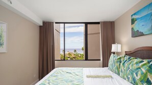 Une chambre d’hôtel avec un lit, une fenêtre avec des rideaux, un tableau et une vue sur l’océan.