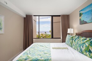Een hotelkamer met een bed, een raam met gordijnen, een schilderij en uitzicht op de oceaan.