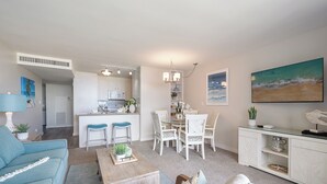 Un salon moderne avec un canapé bleu, une table basse en bois et un coin repas comprenant des chaises blanches, avec une cuisine en arrière-plan.