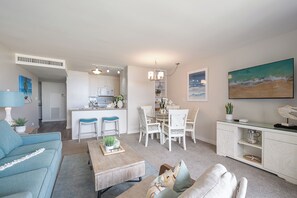 Een moderne woonkamer met een blauwe bank, een houten salontafel, en een eethoek met witte stoelen en een keuken op de achtergrond.