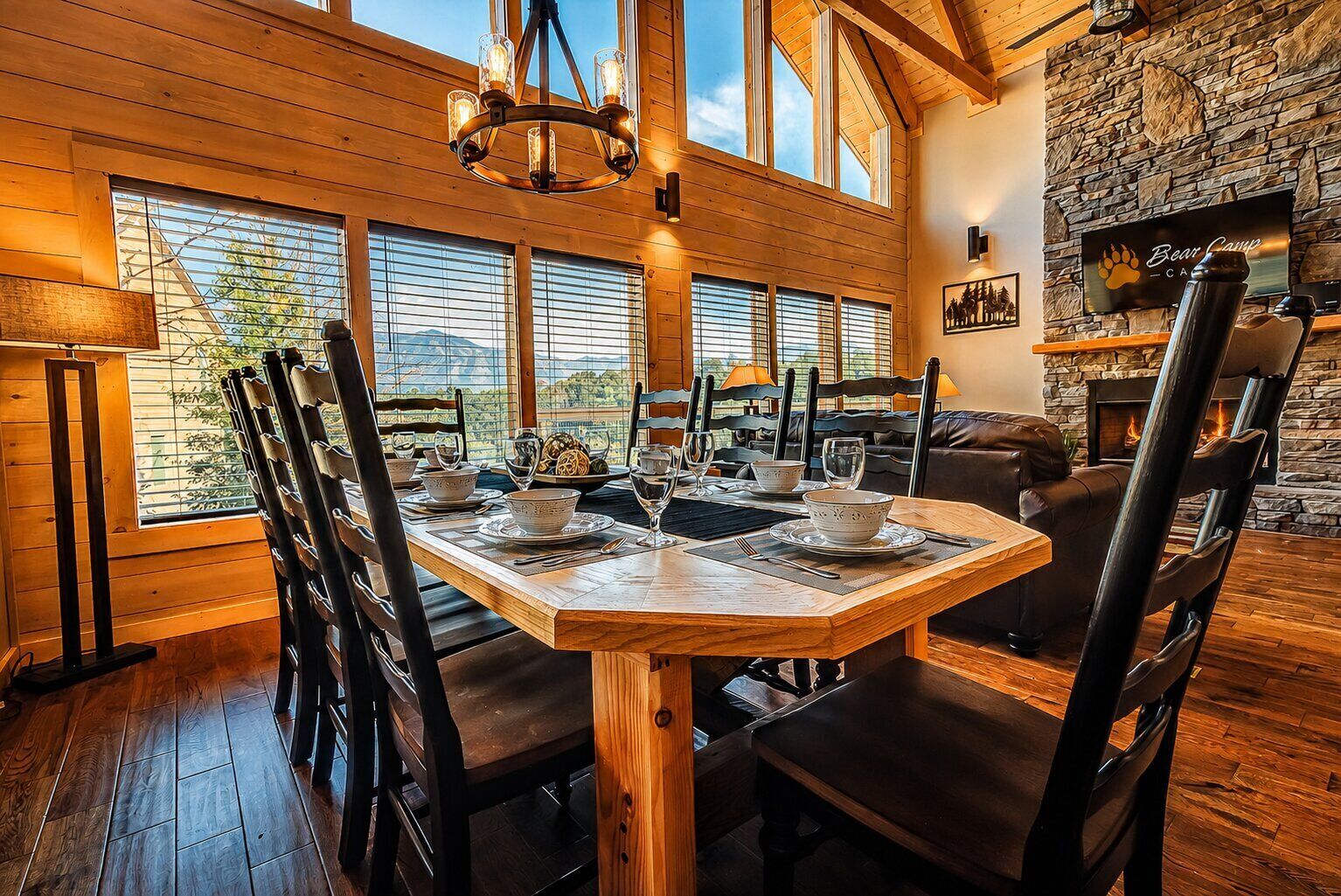 A dining area with a wooden table set for four, surrounded by chairs, with a stone fireplace and a television displaying 'Bear Camp'.