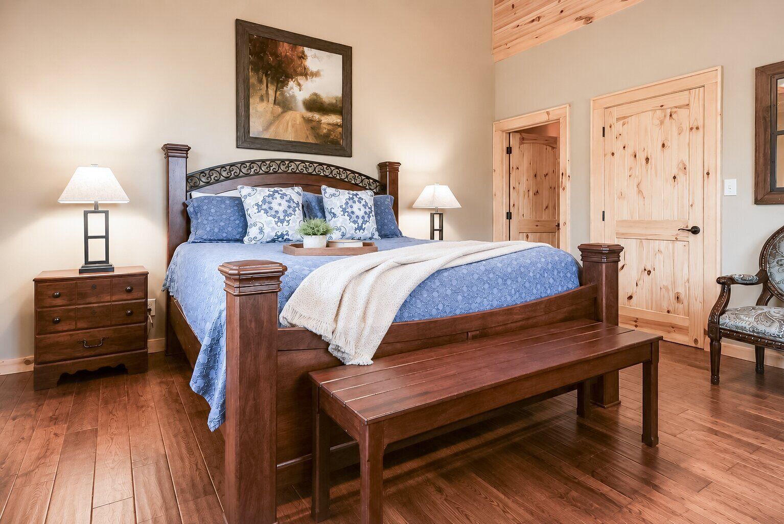 A bedroom with a wooden bed, two bedside tables with lamps, a bench, and a door.