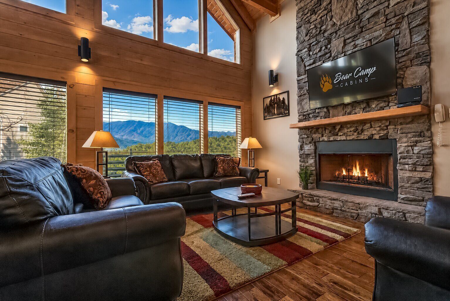 Una sala de estar acogedora con chimenea de piedra, sofás de cuero y un televisor grande mostrando 'Bear Camp'.