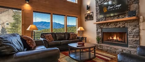 Una sala de estar acogedora con chimenea de piedra, sofás de cuero y un televisor grande mostrando 'Bear Camp'.