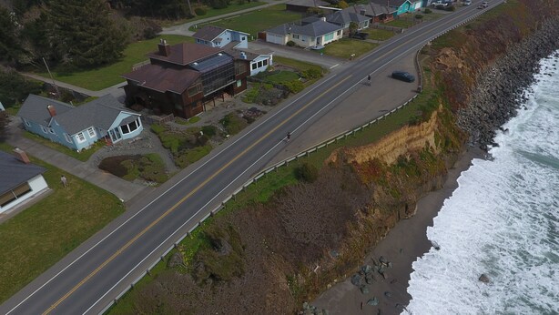 A coastal road with houses on one side and a rocky shoreline on the other.
