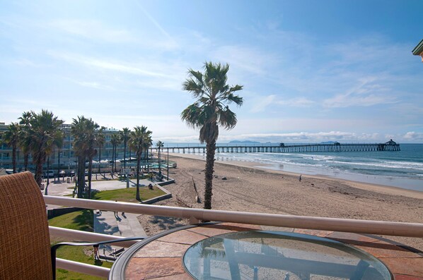 A beach view from a balcony with palm trees, a pier, and a hotel in the background.